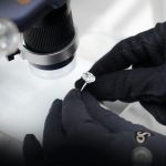 Close-up of a jeweler examining a diamond ring with a microscope and gloved hands.